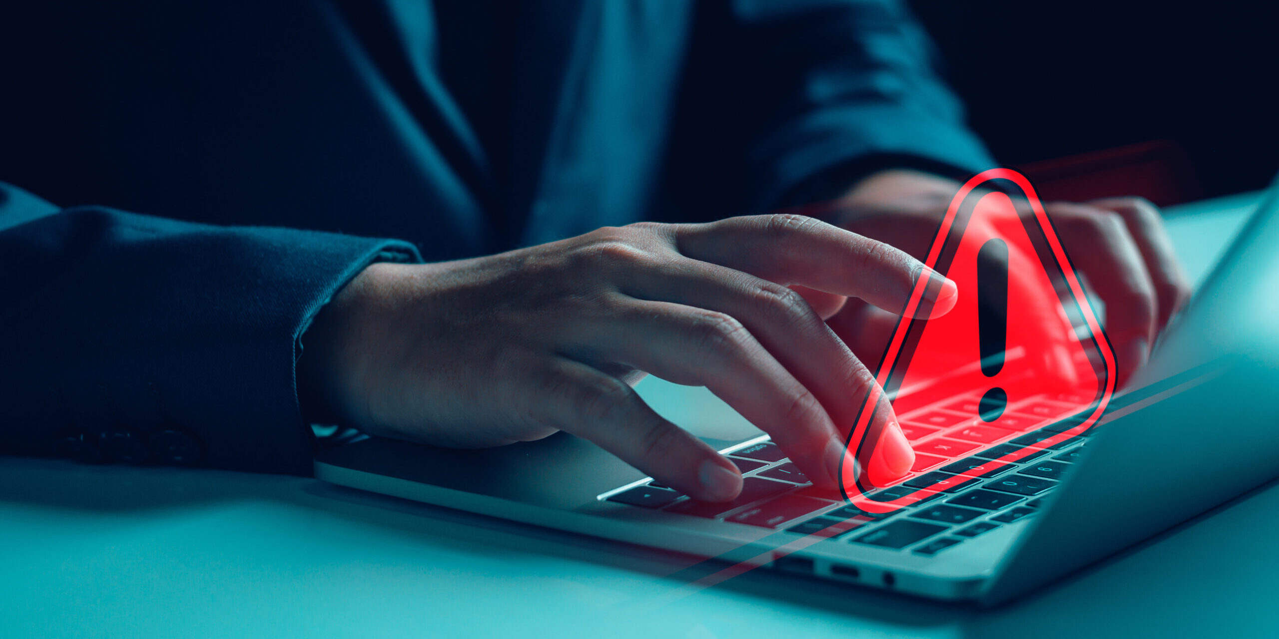 A close-up of a person's hands typing on a laptop with a red warning symbol overlay, representing a cybersecurity threat.