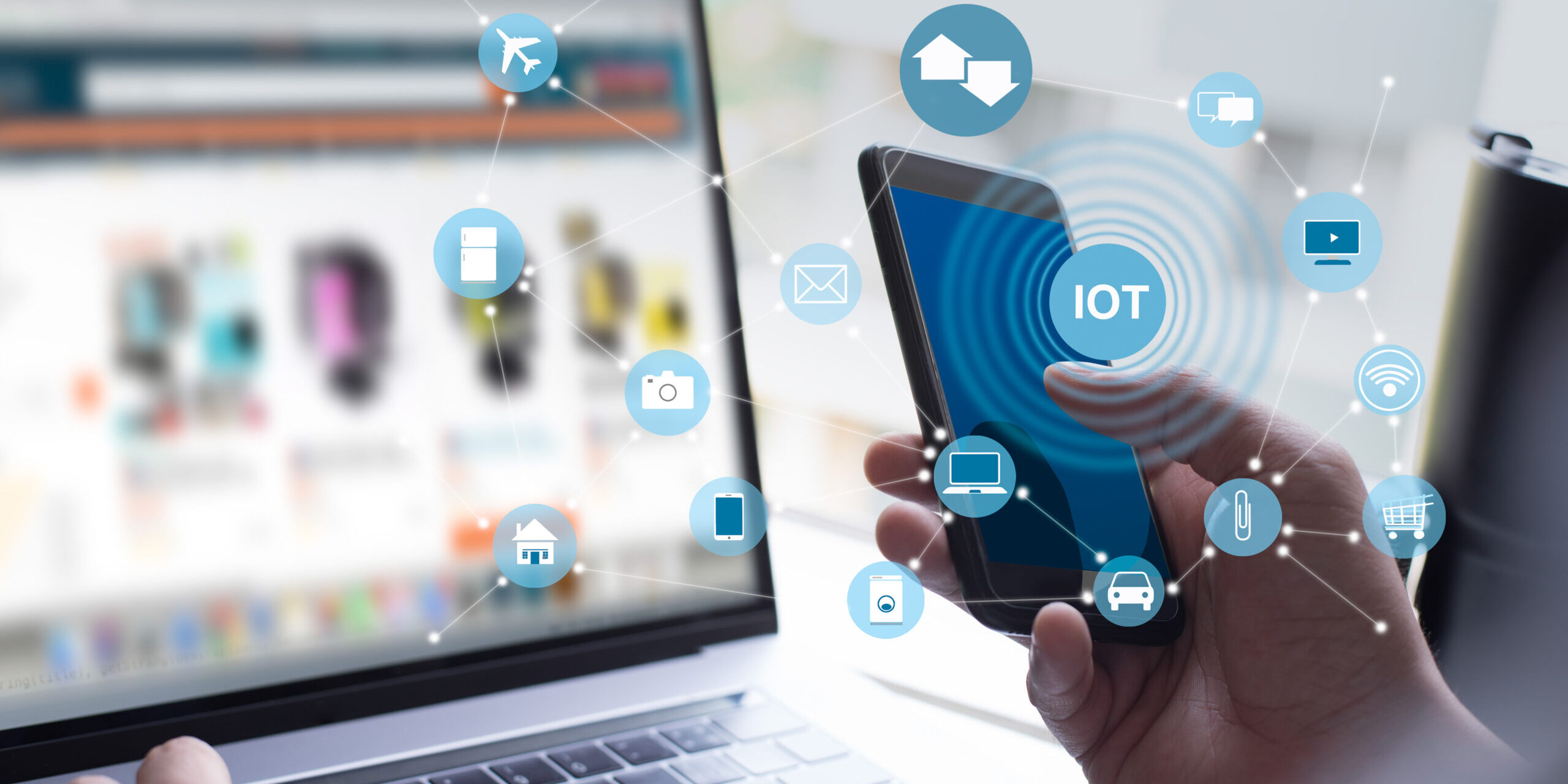 A person holding a smartphone displaying "IoT" while using a laptop, with various interconnected IoT icons representing smart devices such as home automation, security cameras, and smart appliances.