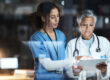 Two healthcare professionals reviewing patient information on a tablet, demonstrating the use of healthcare technology and electronic health records in a clinical environment.