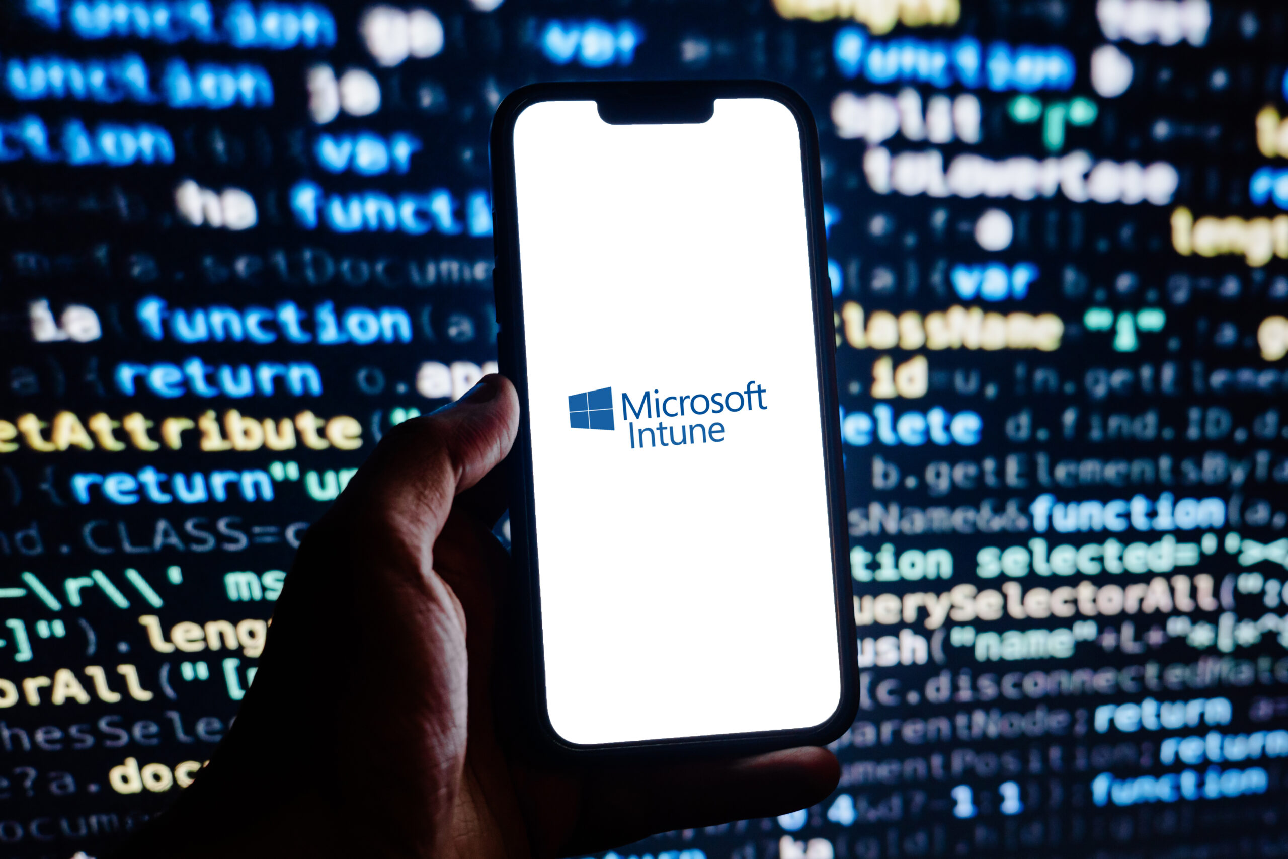 A smartphone displaying the Microsoft Intune logo against a blurred background of computer code, representing cloud-based endpoint management, mobile device security, and Microsoft 365 device administration.